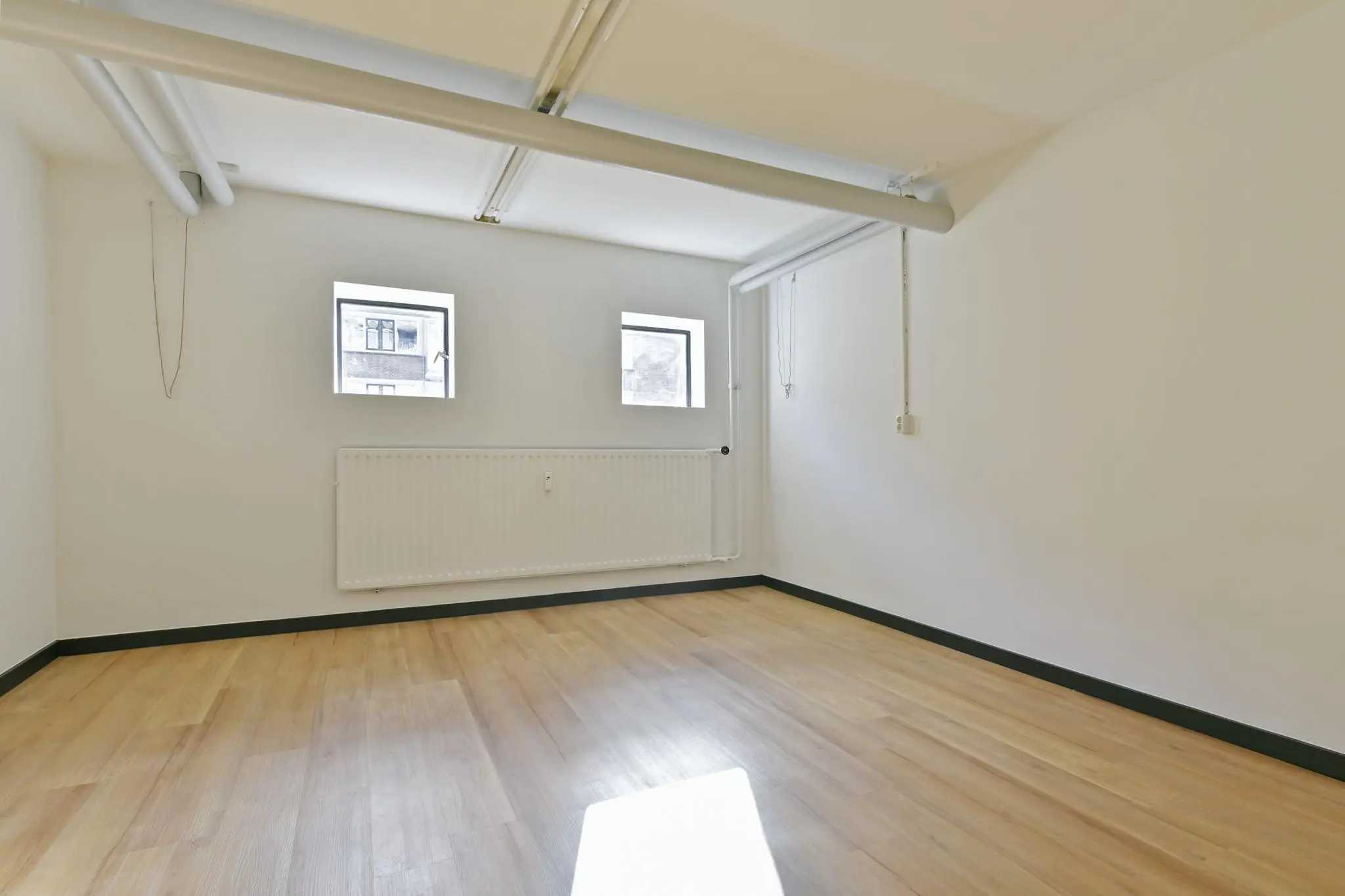 Lege kamer met houten vloer, witte muren, drie kleine ramen en zichtbare leidingen aan het plafond aan de Cornelis Dopperkade.